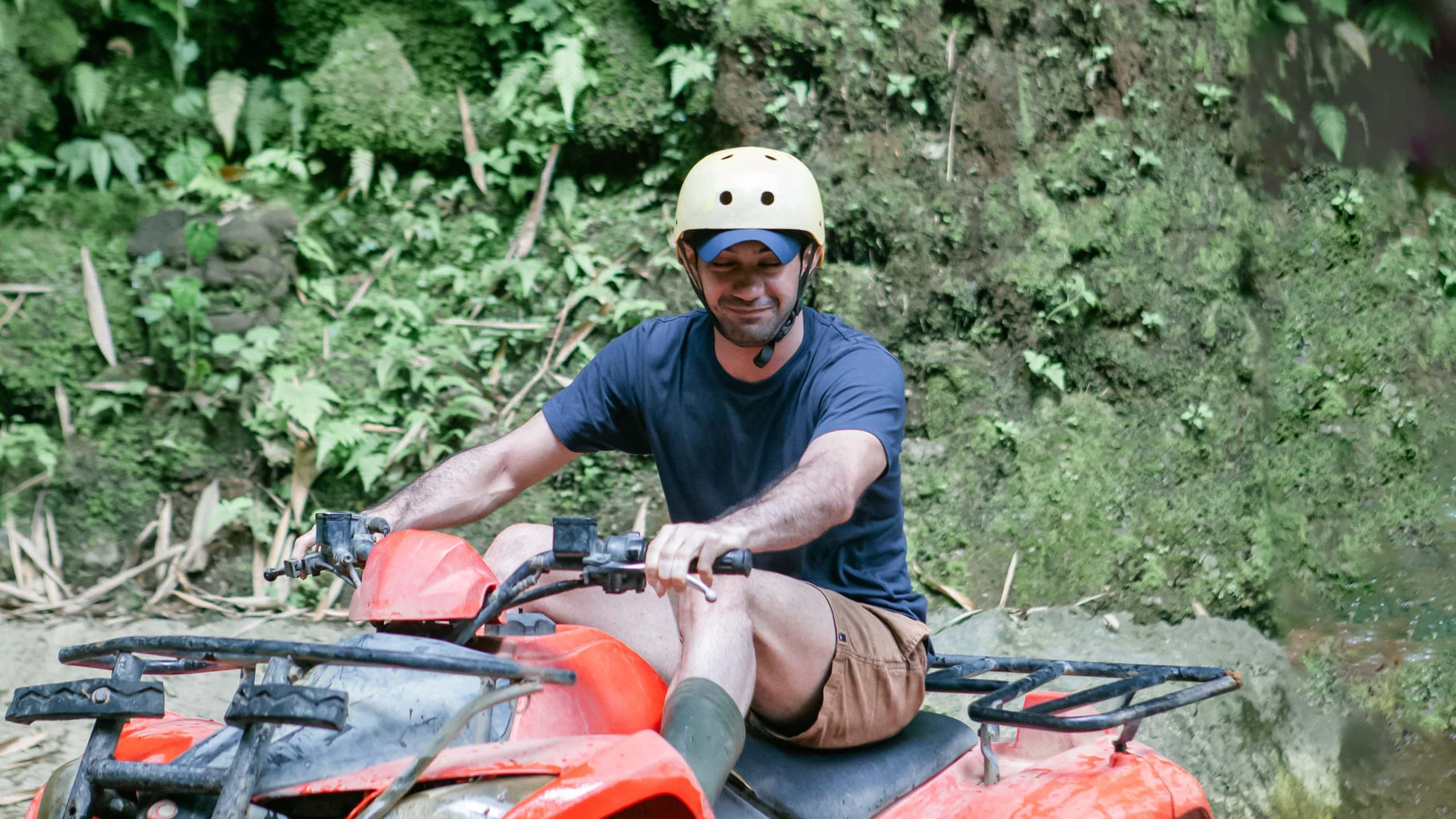 A rider on a Gosek Adventure quad bike crossing a shallow river below a fern-covered jungle wall in Gianyar, Bali