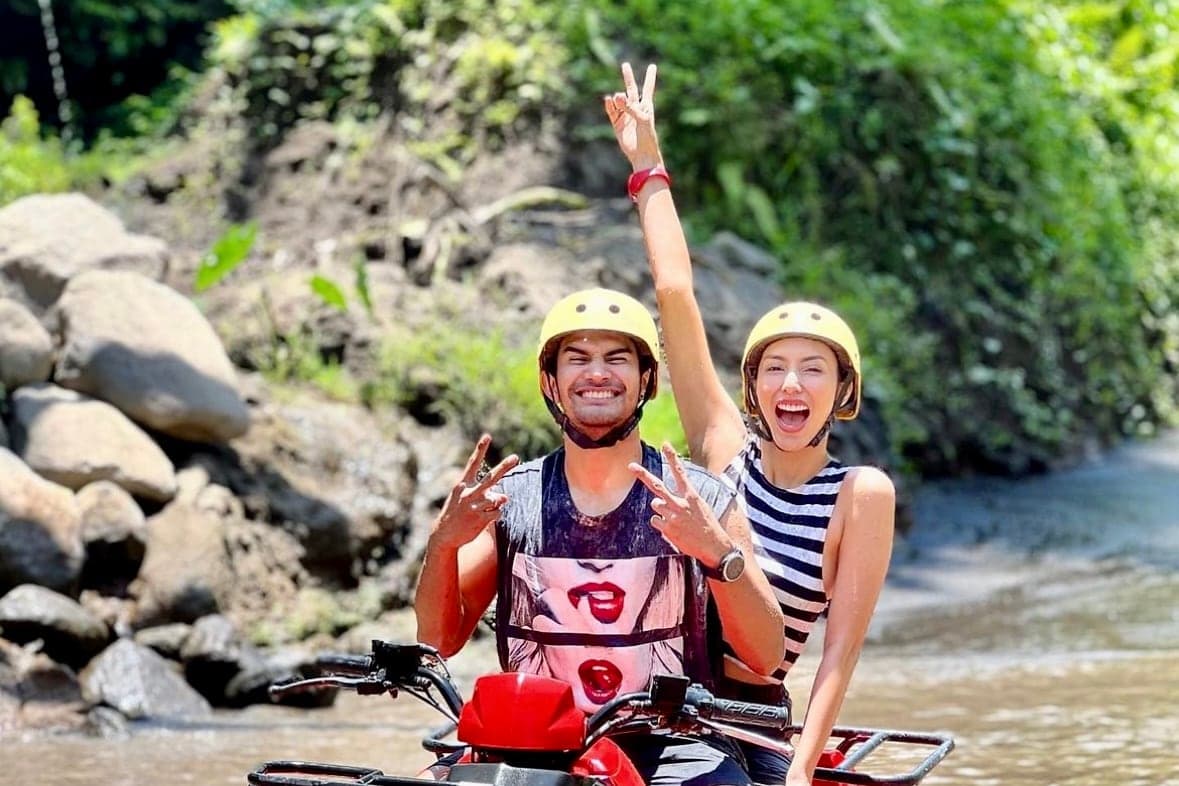 Two riders sharing one red Gosek Adventure tandem quad bike crossing a river, jungle behind them, in Gianyar, Bali